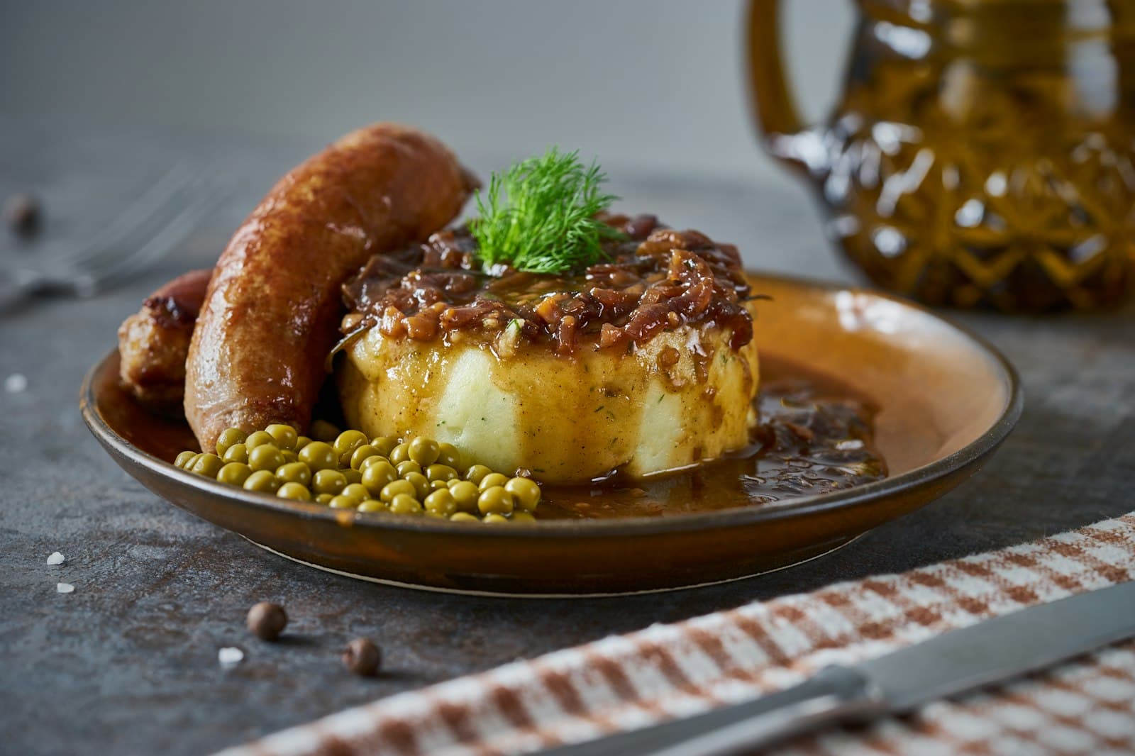 Pork sausages beside a round of mashed potato topped with onion gravy and dill, with green peas on a brown plate
