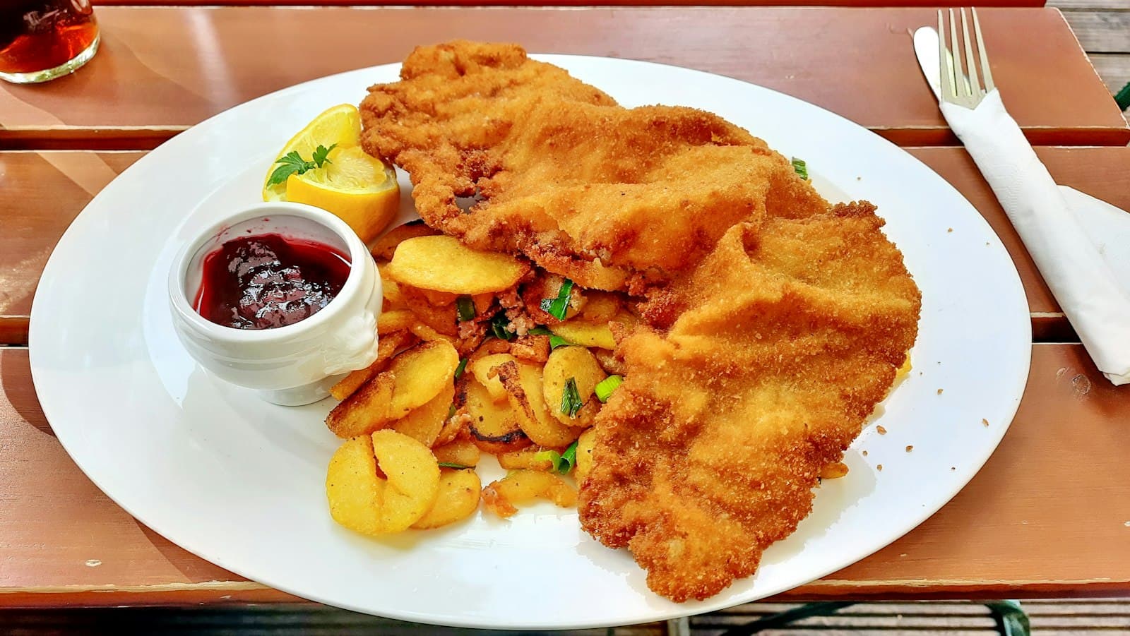 Two breaded pork schnitzels on a white plate with fried potatoes, a lemon wedge and a small dish of red berry sauce