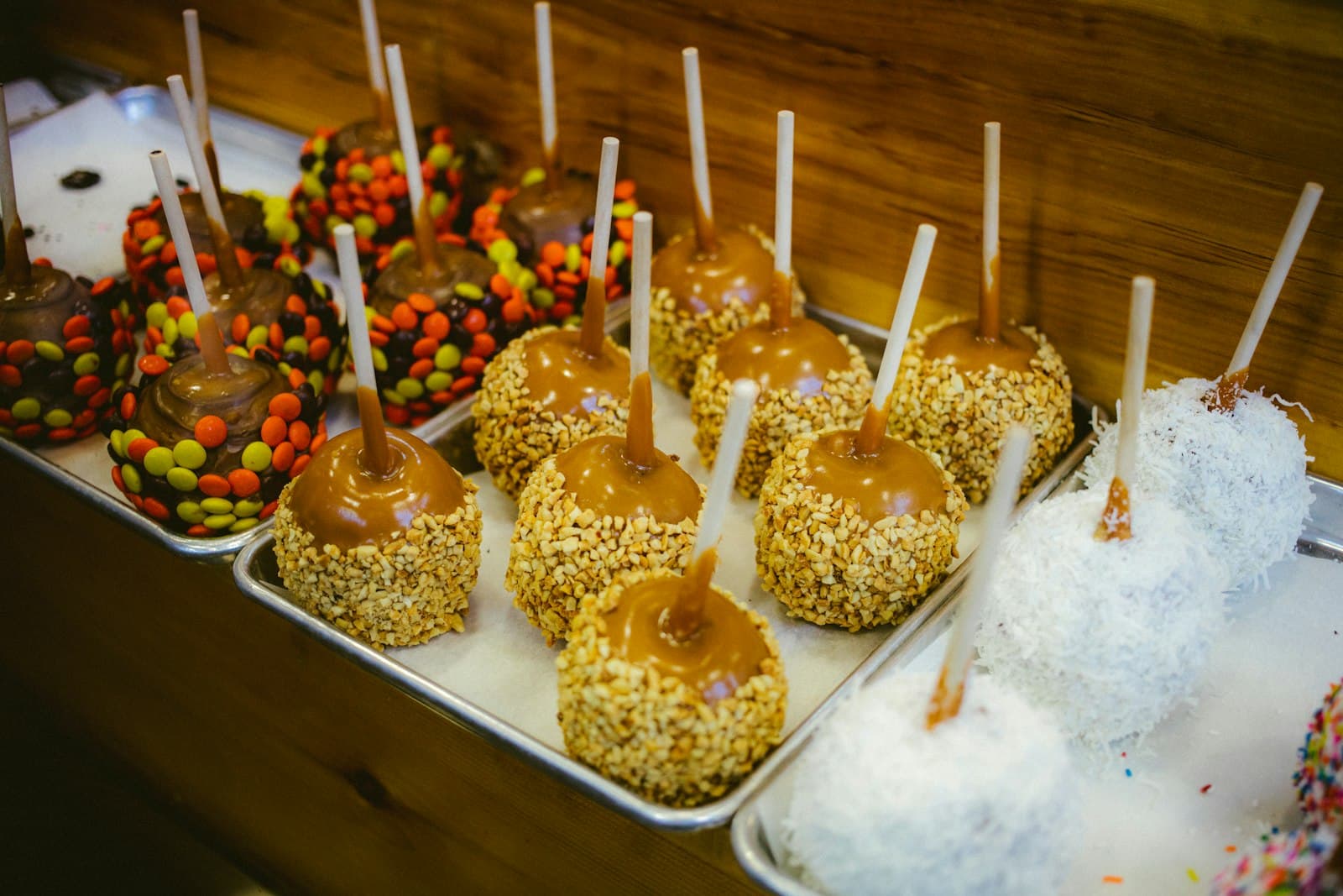 Caramel apples on sticks lined up on metal trays, some rolled in chopped nuts, candy pieces or shredded coconut