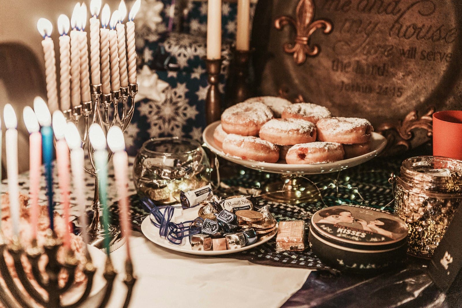 Sugar-dusted jam doughnuts on a cake stand beside lit Hanukkah menorahs and wrapped chocolates