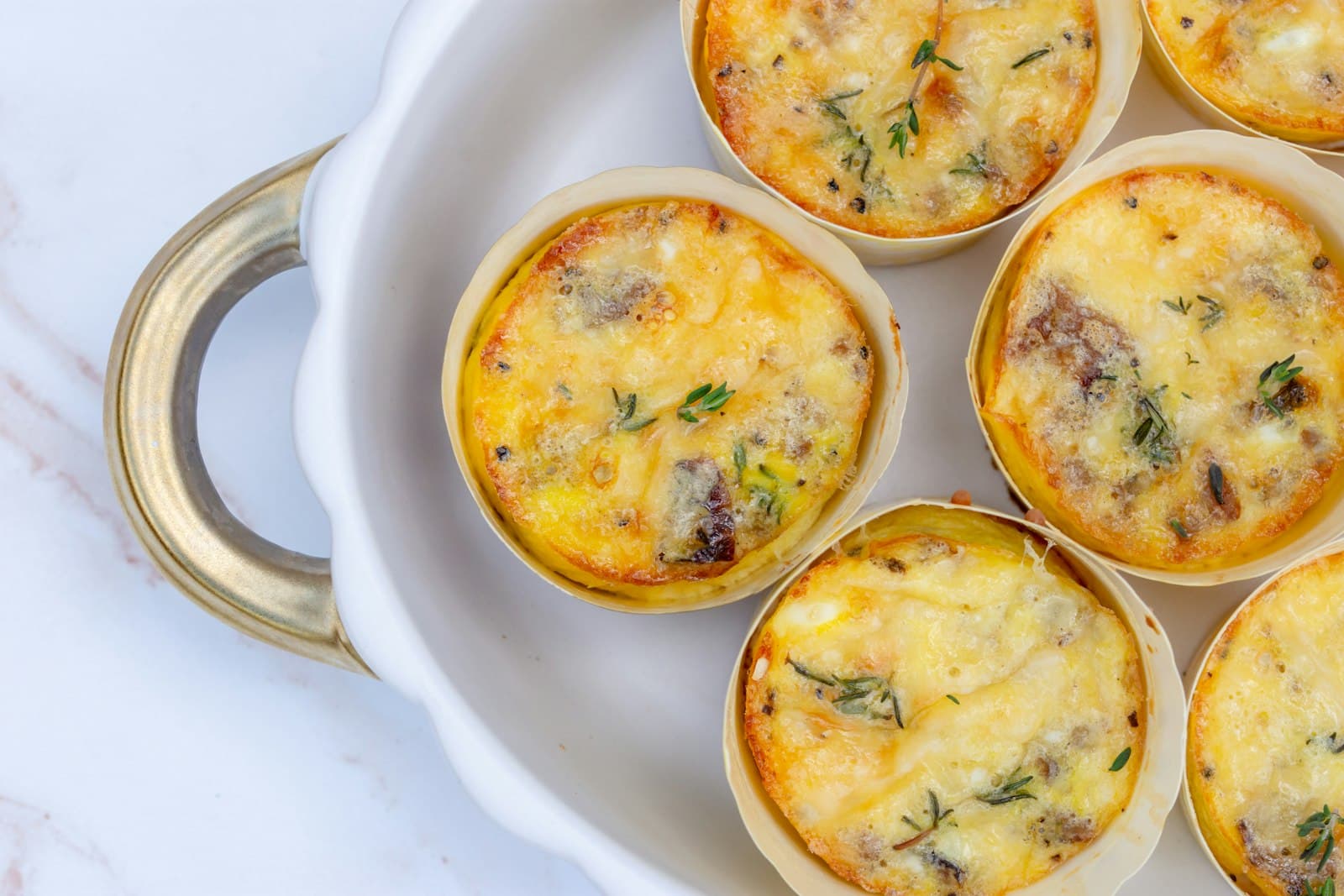 Golden baked egg muffins with melted cheese and fresh thyme in parchment liners inside a white dish