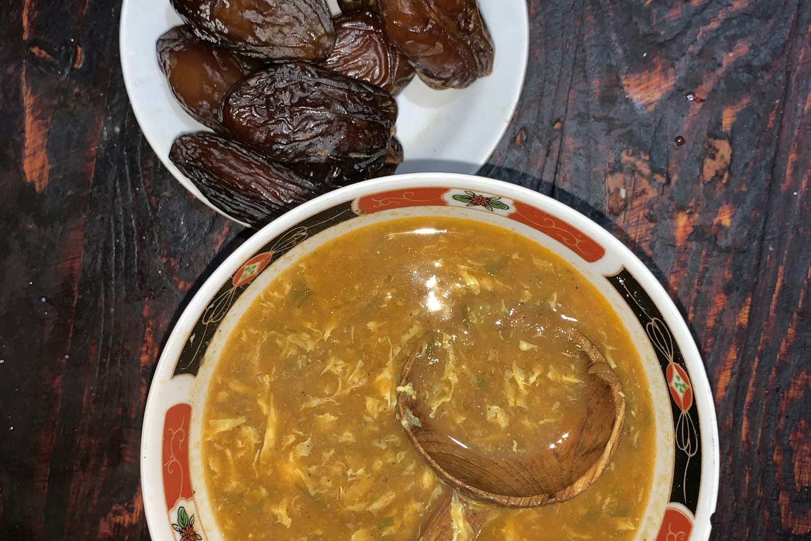 A bowl of thick tomato soup with a wooden spoon, next to a plate of dates on a dark wooden table