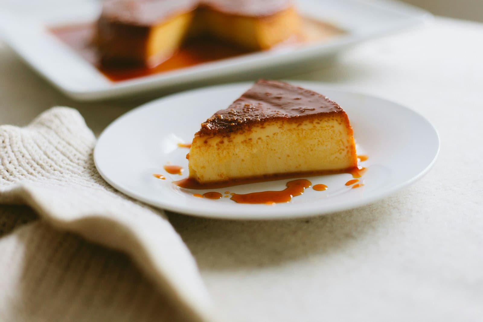 Slice of dense caramel custard flan with dark caramel sauce on a white plate, with the rest of the flan behind