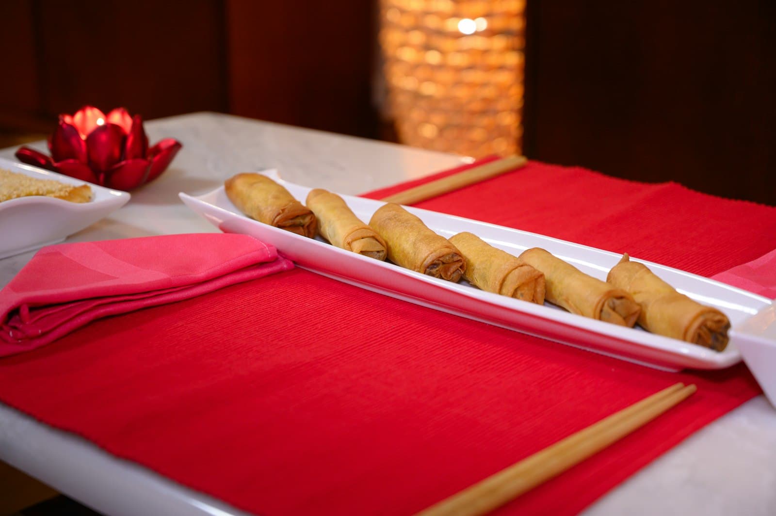 Golden fried spring rolls cut into short pieces on a long white platter, set on a red place mat with chopsticks